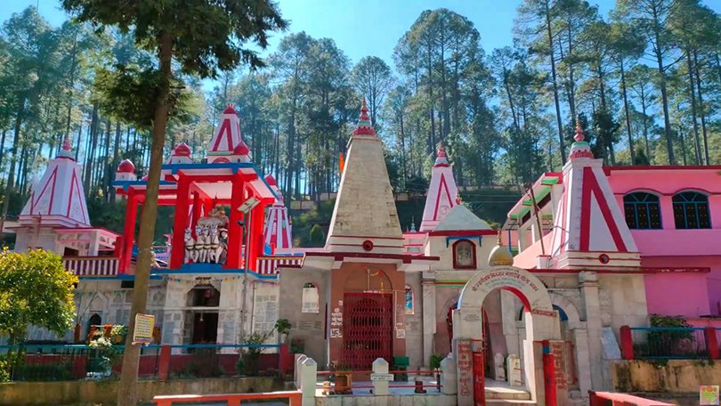 “Ancient stone Binsar Mahadev Temple surrounded by tall pine and cedar trees near Ranikhet, Uttarakhand, with devotees visiting and prayer flags adding color to the peaceful mountain setting.”