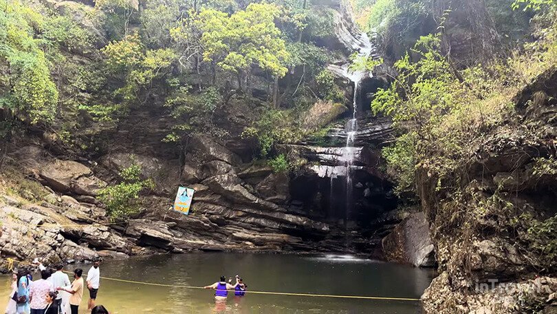 Bhalu Gaad Waterfall flowing into a natural rock pool surrounded by green forest, people are enjoying.